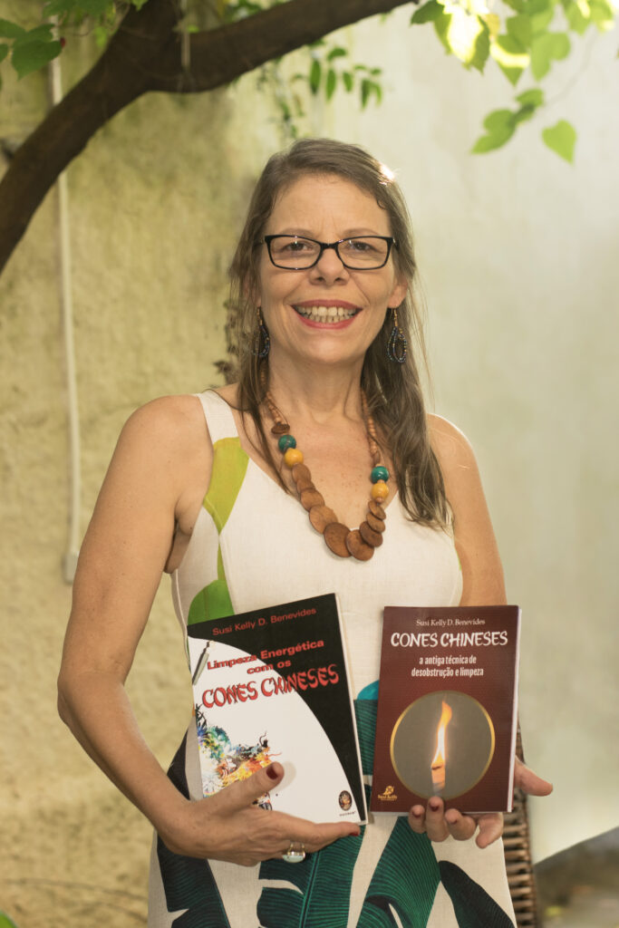 Susi Kelly de óculos, sorridente, segura seus dois livros sobre Cones Chineses aplicado em ouvido para o respiratório e o livro Limpeza energética Cones Chineses